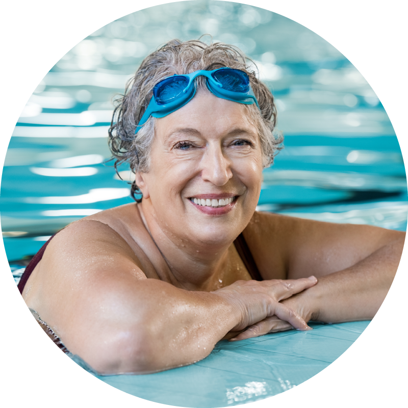 A senior woman in a swimming pool with her arms on the side of the pool looking happy